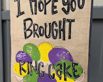 Pode incluir: Uma bandeira de jardim retangular de serapilheira com o texto "I Hope You Brought King Cake" em preto. A bandeira apresenta uma ilustração colorida de bolo rei com cobertura verde, amarela e roxa.
