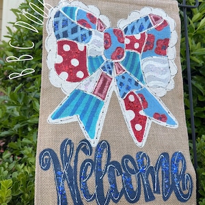 May include: A burlap garden flag with a large bow design. The bow is a patchwork of red, white, and blue patterns. The word "Welcome" is written in blue script with glitter accents. The flag is displayed outdoors.
