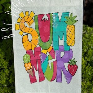 May include: A colorful garden flag with the word "SUMMER" in large, whimsical letters. The design includes a sun, pineapple, ice cream cone, and strawberry. The flag is made of a light-colored fabric and is displayed on a black metal stand.