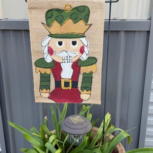 May include: A burlap garden flag featuring a colorful illustration of a nutcracker soldier. The nutcracker wears a green and gold crown, a red jacket, and a white mustache. The flag is displayed on a black metal stand, with a solar-powered light in the foreground.