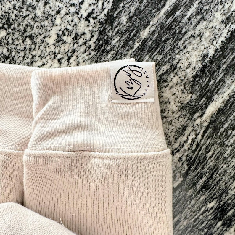 May include: Close-up of light pink fabric with a ribbed cuff. A white tag is sewn on the cuff with the black text "Kozy Apparel" and a circular logo. The fabric appears to be soft and comfortable.