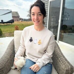 May include: Beige sweatshirt featuring embroidered chicks in black, white, and yellow. The sweatshirt is worn by a person sitting outdoors. The person is wearing blue jeans and is holding a fluffy white chicken.
