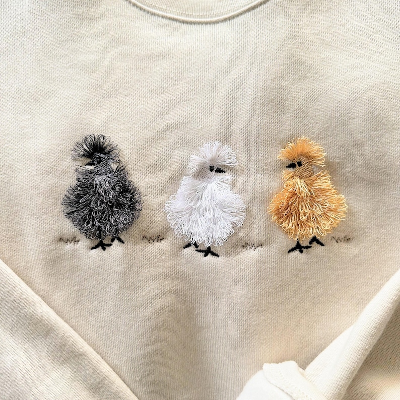 May include: Cream-colored sweatshirt featuring three embroidered chicks in a row. The chicks are black, white, and golden yellow, with fluffy textures and black feet. The design is simple and whimsical, perfect for casual wear.