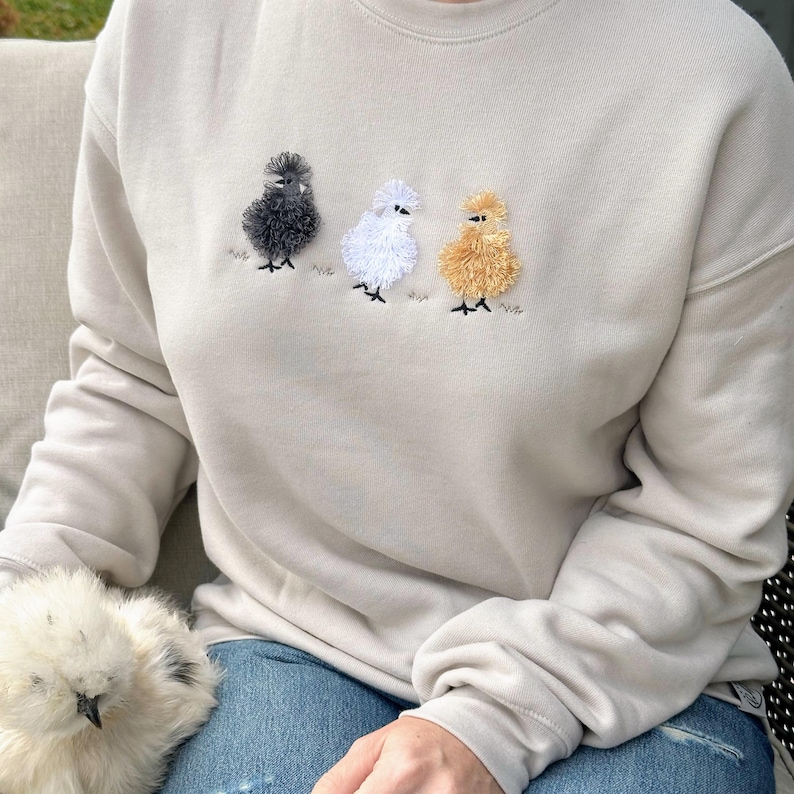 May include: Beige sweatshirt featuring embroidered chickens in black, white, and gold. The chickens are standing on a patch of green grass. The sweatshirt is a neutral color and has long sleeves.