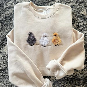 May include: Cream-colored sweatshirt with embroidered design of three fluffy chickens in gray, white, and gold. The chickens are standing on a line of embroidered grass. The sweatshirt has a crew neck and long sleeves.