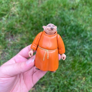 May include: A small plastic figurine of a bear character wearing a bright orange monk-style robe with a thin, light-colored rope belt tied around the waist. The bear has a light tan face, small rounded ears, and a gentle, smiling facial expression.