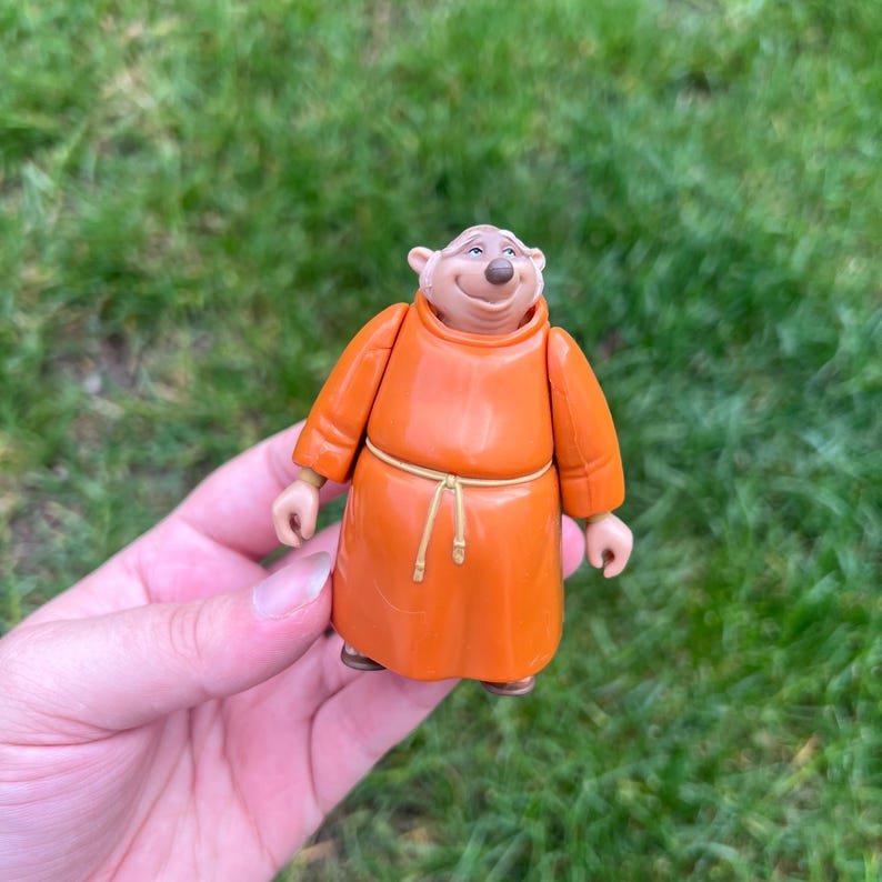May include: A small plastic figurine of a bear character wearing a bright orange monk-style robe with a thin, light-colored rope belt tied around the waist. The bear has a light tan face, small rounded ears, and a gentle, smiling facial expression.