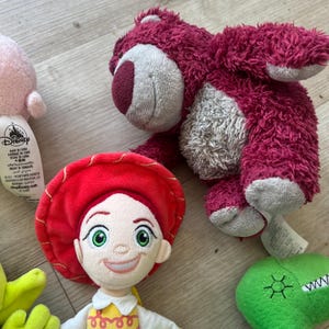 K&ouml;nnte beinhalten: Eine Sammlung von Disney-Pl&uuml;schtieren, darunter eine rot-wei&szlig;e Jessie-Puppe, ein rosa B&auml;r und ein gr&uuml;ner Dinosaurier. Die Spielzeuge bestehen aus weichen Materialien und weisen detaillierte Gesichtsausdr&uuml;cke auf.