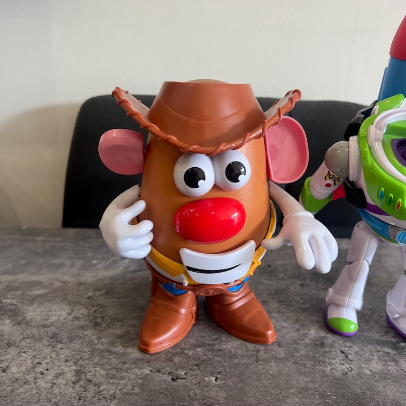 May include: A toy Mr. Potato Head figure dressed as a cowboy, with a brown hat, boots, and a red nose. The figure has large eyes and white gloves. A Buzz Lightyear toy is partially visible on the right.