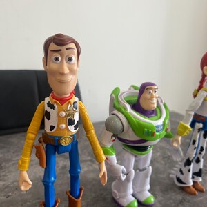 K&ouml;nnte beinhalten: Drei Kunststoff-Actionfiguren von Charakteren aus dem Animationsfilm Toy Story. Woody, in Cowboy-Outfit, steht neben Buzz Lightyear in einem gr&uuml;n-wei&szlig;en Raumanzug. Eine dritte Figur, Jessie, ist teilweise sichtbar.
