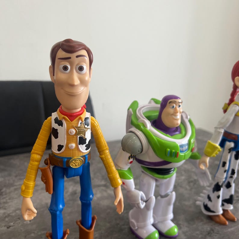 K&ouml;nnte beinhalten: Drei Kunststoff-Actionfiguren von Charakteren aus dem Animationsfilm Toy Story. Woody, in Cowboy-Outfit, steht neben Buzz Lightyear in einem gr&uuml;n-wei&szlig;en Raumanzug. Eine dritte Figur, Jessie, ist teilweise sichtbar.