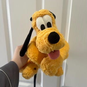 May include: A plush toy of Pluto, a yellow dog with black ears and nose, and a pink tongue sticking out. The toy has large white eyes with black pupils. The plush is soft and fluffy, with a friendly expression.