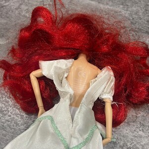Puede incluir: Una mu&ntilde;eca de pelo rojo con un vestido blanco con ribetes verdes. La espalda de la mu&ntilde;eca es visible y el vestido est&aacute; abierto por la espalda. La mu&ntilde;eca tiene una marca CE en la espalda.