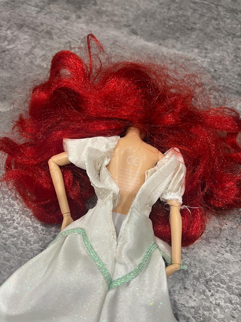 Puede incluir: Una mu&ntilde;eca de pelo rojo con un vestido blanco con ribetes verdes. La espalda de la mu&ntilde;eca es visible y el vestido est&aacute; abierto por la espalda. La mu&ntilde;eca tiene una marca CE en la espalda.