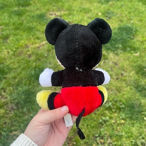May include: A small Mickey Mouse plush toy, viewed from the back, with black ears, a black head, and a red body. The toy has white gloves, yellow feet, and a black tail. The plush is held against a background of green grass.
