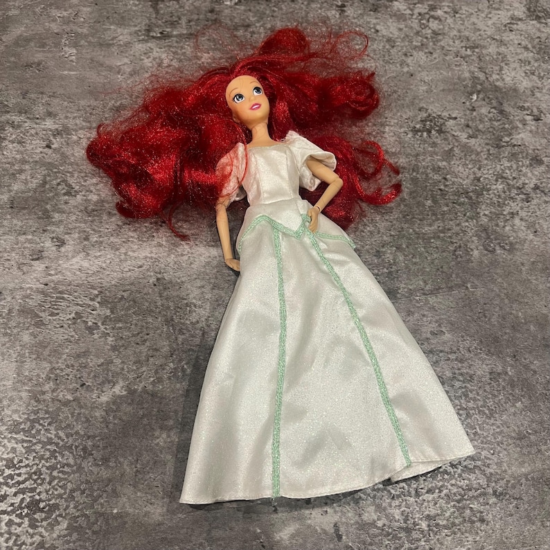 Puede incluir: Una mu&ntilde;eca de pelo rojo con un vestido blanco con ribete verde. La mu&ntilde;eca tiene una larga peluca roja y lleva un vestido blanco con ribete verde. La mu&ntilde;eca est&aacute; mirando hacia arriba.