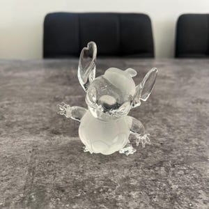 May include: A frosted glass figurine of Stitch from Lilo & Stitch wearing a hat. The figurine is white and has a detailed design.