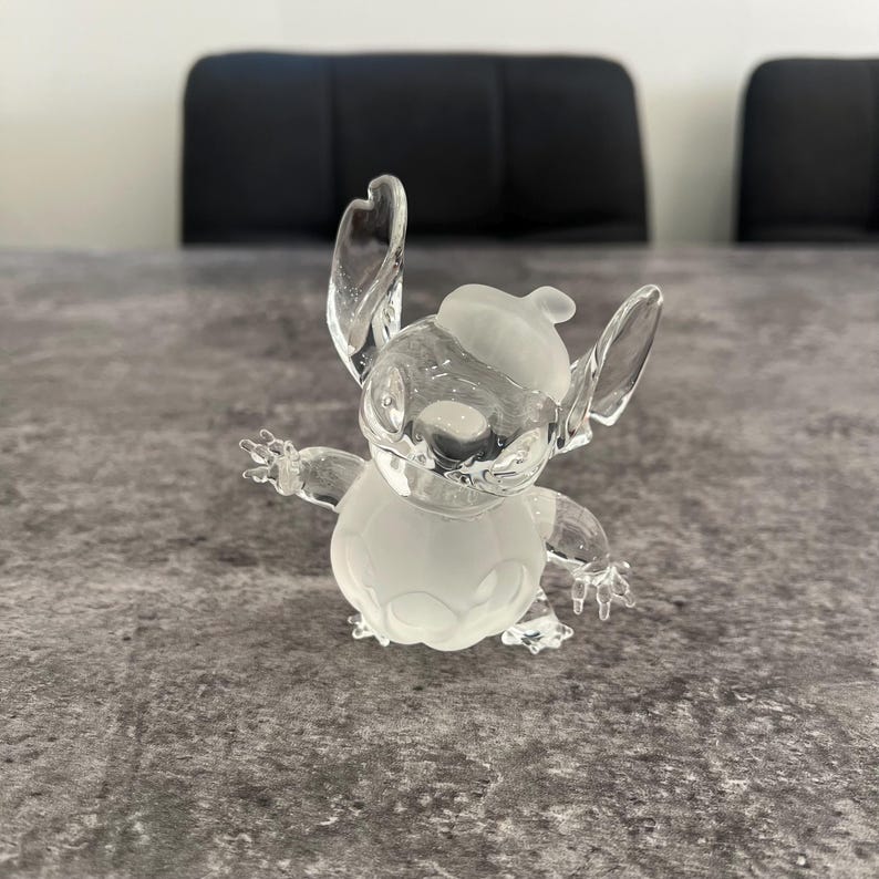 May include: A frosted glass figurine of Stitch from Lilo & Stitch wearing a hat. The figurine is white and has a detailed design.