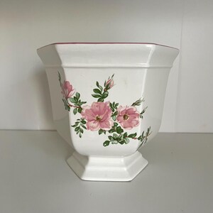 May include: White ceramic planter with a hexagonal shape and a pedestal base. The planter is decorated with pink floral designs and green leaves. The rim is accented with a thin pink line. A decorative piece for indoor or outdoor use.