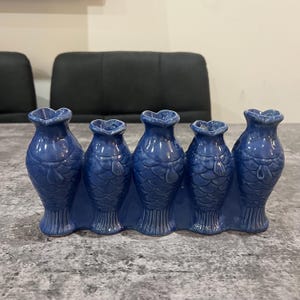 May include: A blue ceramic centerpiece featuring five fish-shaped vases. Each vase has a heart-shaped opening and is connected at the base. The fish scales are detailed, and the vases are arranged in a row, creating a decorative display.