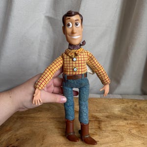 May include: A plastic and fabric doll featuring a molded brown hair head, a yellow long-sleeved shirt with a red grid pattern, a brown neckerchief, blue denim-style trousers with a brown belt, and brown cowboy boots with silver-colored accents.