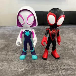 May include: Two action figures of Spider-Man characters. One is a white figure with pink and blue accents, and the other is a black figure with red accents.