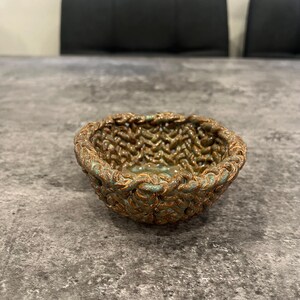 May include: A small, handcrafted ceramic bowl with a textured, woven-like design. The bowl features a blend of bronze and teal colors, creating a unique and artistic appearance. The interior has a teal glaze.