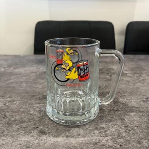 May include: Clear glass beer mug with a handle. The mug features a cartoon image of Homer Simpson with the words "Duff Beer" and "Can't get enough..." printed on the front. The mug is sitting on a gray surface.