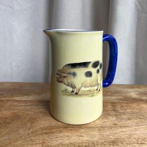 May include: A cream-colored ceramic pitcher with a blue handle and rim. The pitcher features a detailed illustration of a pig with black spots. The pig is standing on a patch of grass. The pitcher is on a wooden surface.
