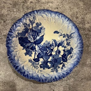 May include: A round, antique ceramic plate with a blue and white floral design. The plate has a raised floral pattern in the centre and a scalloped edge. The flowers are a deep blue against a cream background.