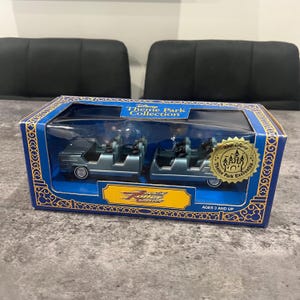May include: A boxed Disney Theme Park Collection toy set featuring two miniature roller coaster cars. The cars are blue-grey with clear seating. The box is blue with gold accents and the text "Disney Theme Park Collection" and "Ages 3 and Up".