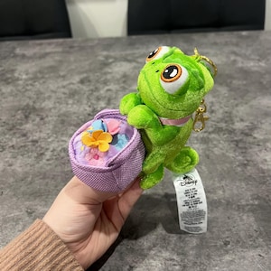 May include: A green plush chameleon keyring holding a small purple basket with fabric flowers. The chameleon has large eyes and a pink collar. A Disney tag is attached.