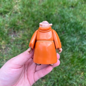 May include: A small plastic figurine of a monk wearing a solid orange robe with a light-colored cord tied around the waist. The figure has a bald head with a ring of hair and is shown from the back, displaying the smooth texture of the garment.