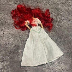 Puede incluir: Una mu&ntilde;eca de pelo rojo con un vestido blanco con ribete verde. La mu&ntilde;eca est&aacute; tumbada sobre una superficie gris.