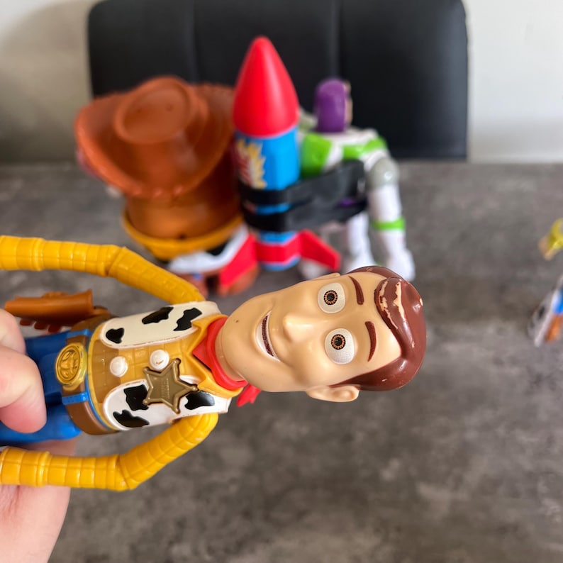May include: A close-up of a Woody action figure with a smiling face, wearing a cowboy hat, vest, and red neckerchief. A red, blue, and white rocket, and a Buzz Lightyear figure are in the background.