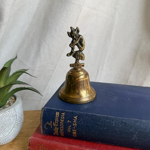 May include: A brass bell featuring a small figure of a seated imp or goblin on top. The base of the handle is inscribed with the word DEVON. The bell has a polished metallic finish and is positioned on top of a blue hardbound book.