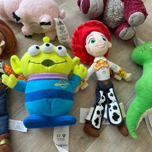 K&ouml;nnte beinhalten: Eine Sammlung farbenfroher Pl&uuml;schtiere mit Charakteren aus dem Toy Story-Franchise. Die Spielzeuge umfassen einen gr&uuml;nen, dre&auml;ugigen Alien, Jessie das Cowgirl, ein rosa Schwein und einen gr&uuml;nen Dinosaurier. Die Spielzeuge sind aus weichen Materialien gefertigt und haben detaillierte Merkmale.