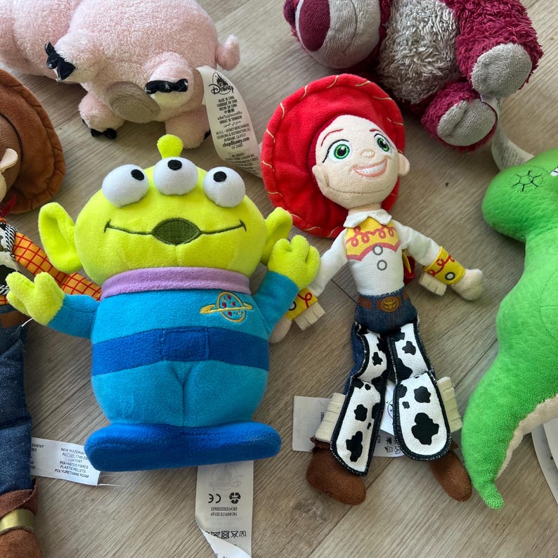K&ouml;nnte beinhalten: Eine Sammlung farbenfroher Pl&uuml;schtiere mit Charakteren aus dem Toy Story-Franchise. Die Spielzeuge umfassen einen gr&uuml;nen, dre&auml;ugigen Alien, Jessie das Cowgirl, ein rosa Schwein und einen gr&uuml;nen Dinosaurier. Die Spielzeuge sind aus weichen Materialien gefertigt und haben detaillierte Merkmale.