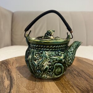 May include: A green ceramic teapot with a black handle and lid. The teapot features a detailed design of seahorses and other sea creatures. The handle is made of a black, coiled material. The teapot is sitting on a wooden surface.
