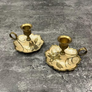 May include: A pair of brass candle holders with a floral design. The holders have a white enamel finish and a small handle on the side.