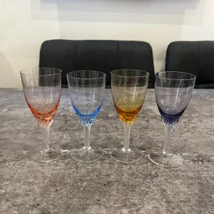 May include: Four coloured wine glasses with clear bowls and twisted stems. The glasses have coloured accents in shades of orange, blue, yellow, and purple. The glasses are arranged on a grey surface.