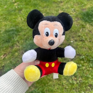 May include: A plush Mickey Mouse toy. The toy features a black body, ears, and head, with a peach-colored face and white hands. It wears red shorts with yellow buttons and yellow shoes. The toy is sitting.