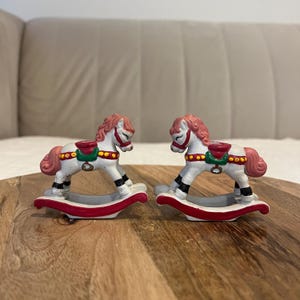 May include: Two miniature rocking horse figurines. The horses are white with pink manes and tails, red saddles, and black hooves. They sit on red and white rockers. The figurines are displayed on a wooden surface.