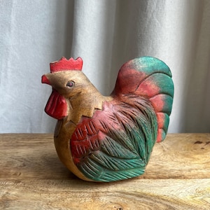 May include: A hand-carved wooden rooster figurine. The rooster features a red comb and wattle, with a body painted in shades of red, green, and brown. The figurine is placed on a wooden surface.