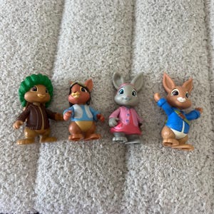 May include: Four small plastic rabbit figurines standing in a row. The first wears a green hat and brown jacket, the second a blue vest and aviator cap, the third a pink coat, and the fourth a blue jacket with a green strap across the chest.