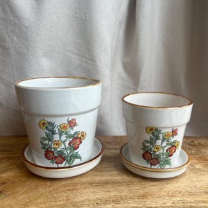 May include: Two ceramic flower pots with matching saucers. The pots are white with a floral design in red, yellow, and green. The rims and saucers have a brown trim. The pots are sitting on a wooden surface.
