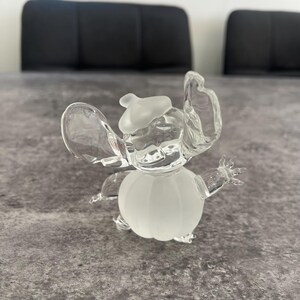 May include: A clear glass figurine of Stitch from Lilo & Stitch. The figurine is wearing a white hat and has a frosted body.