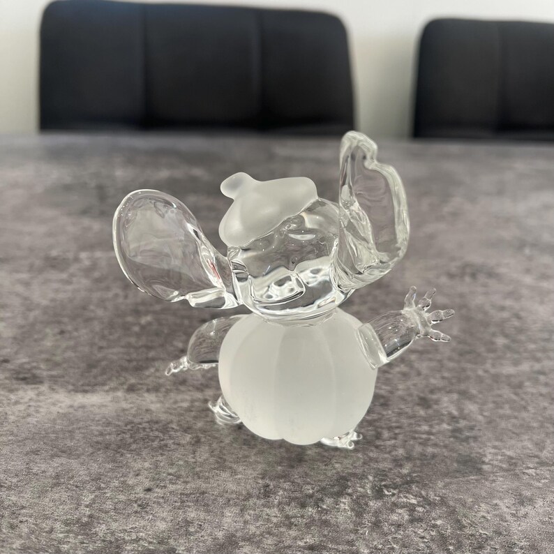 May include: A clear glass figurine of Stitch from Lilo & Stitch. The figurine is wearing a white hat and has a frosted body.