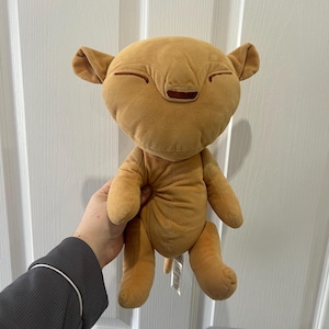 May include: A tan plush toy shaped like a lion cub with a large, rounded head, small ears, and embroidered closed eyes. The soft fabric figure features a simple, minimalist design with a dark brown stitched nose and a smooth, uniform texture.