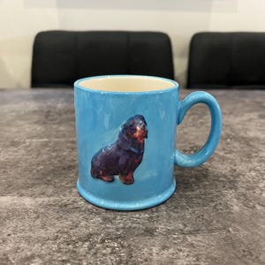 May include: A light blue ceramic mug with a black and brown dog illustration. The mug has a rounded handle and a cream-colored interior. The dog illustration is centered on the mug's exterior.